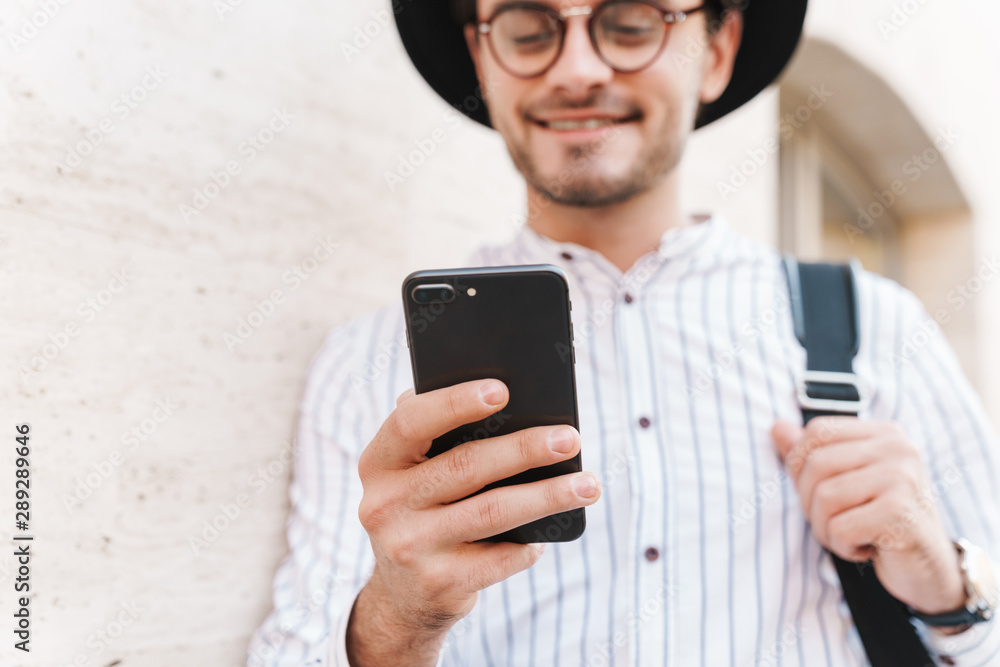 Photo of joyful caucasian man typing on cellphone and smiling while leaning on wall in city street Photo of joyful caucasian man typing on cellphone and smiling while leaning on wall in city street