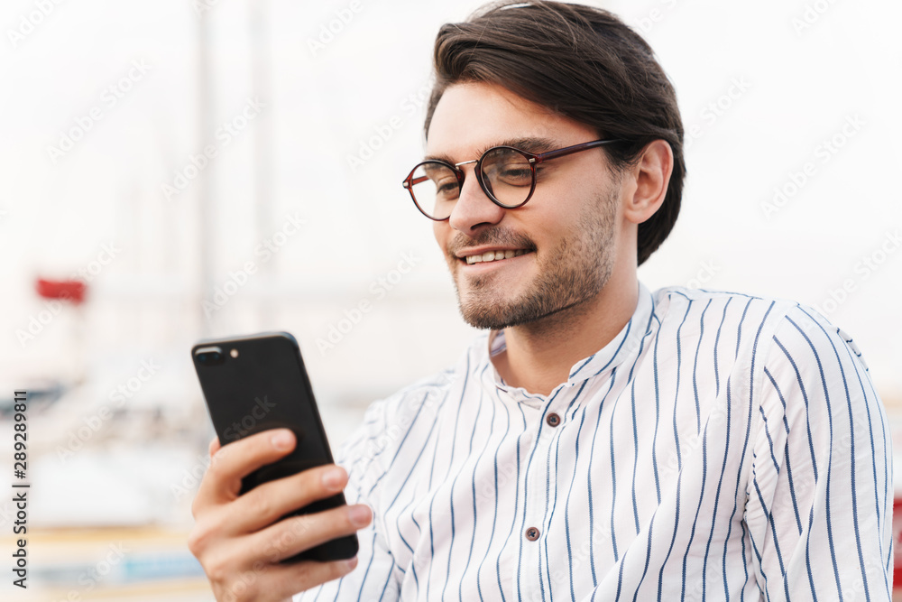 Photo of handsome young man typing on cellphone and smiling while walking on pier Photo of handsome young man typing on cellphone and smiling while walking on pier