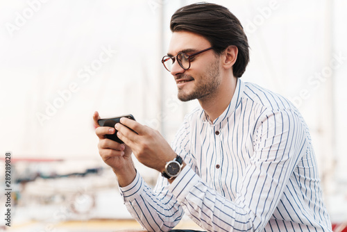 Photo of handsome unshaven man typing on cellphone and smiling while walking on pier