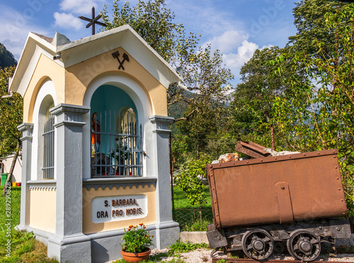 Fotografie Saint Barbara Chapel with mining Vehicle near Graveyard and Mining Tunnel in Log pod Mangartom, Bovec, Slovenia, Europe