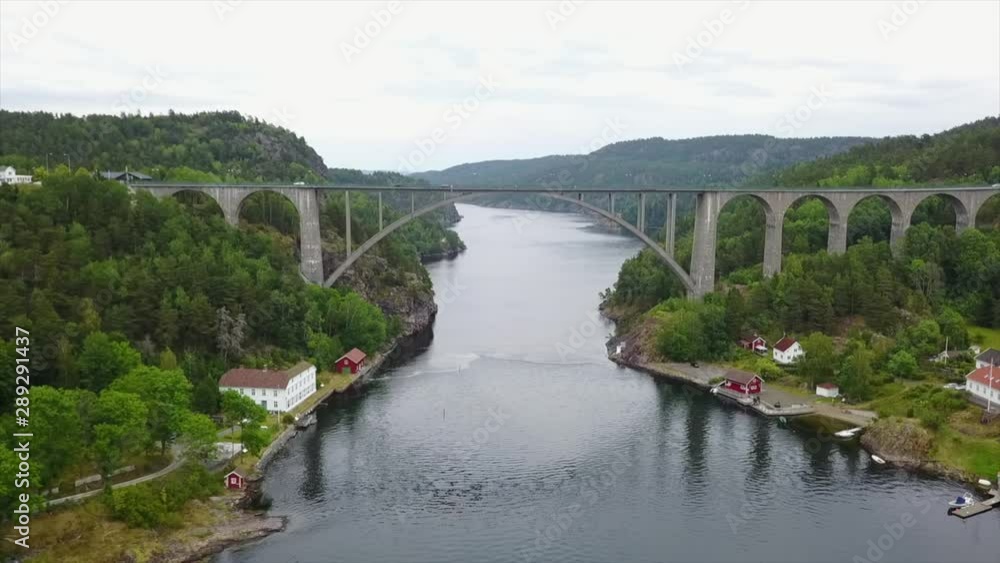 Arched road bridge spanning across narrow watercourse with cars driving, aerial dolly in view of Scandinavian fjord with traditional houses on the shore, travel, construction and transportation Arched road bridge spanning across narrow watercourse with cars driving, aerial dolly in view of Scandinavian fjord with traditional houses on the shore, travel, construction and transportation