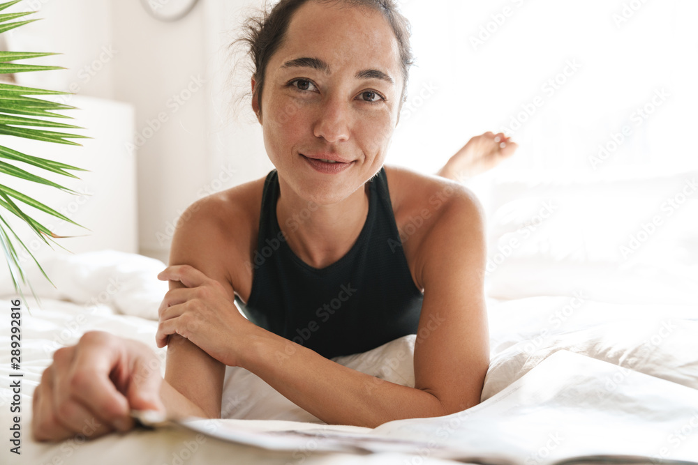 Portrait of slim woman smiling while lying and reading magazine on bed in bright room with green plant Portrait of slim woman smiling while lying and reading magazine on bed in bright room with green plant