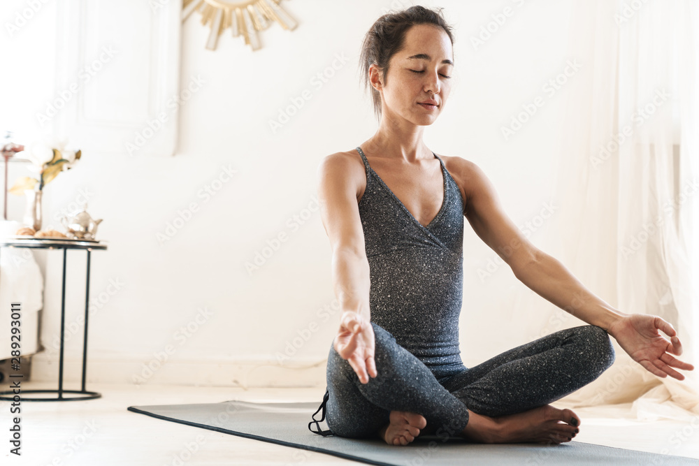 Photo of beautiful sportswoman sitting on fitness mat and meditating while practicing yoga in bright room Photo of beautiful sportswoman sitting on fitness mat and meditating while practicing yoga in bright room