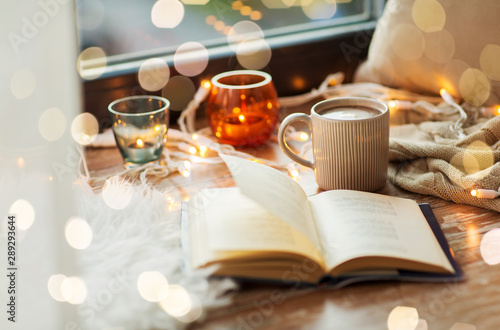 hygge and cozy home concept - book, cup of coffee or hot cchocolate and candles with garland on window sill photo