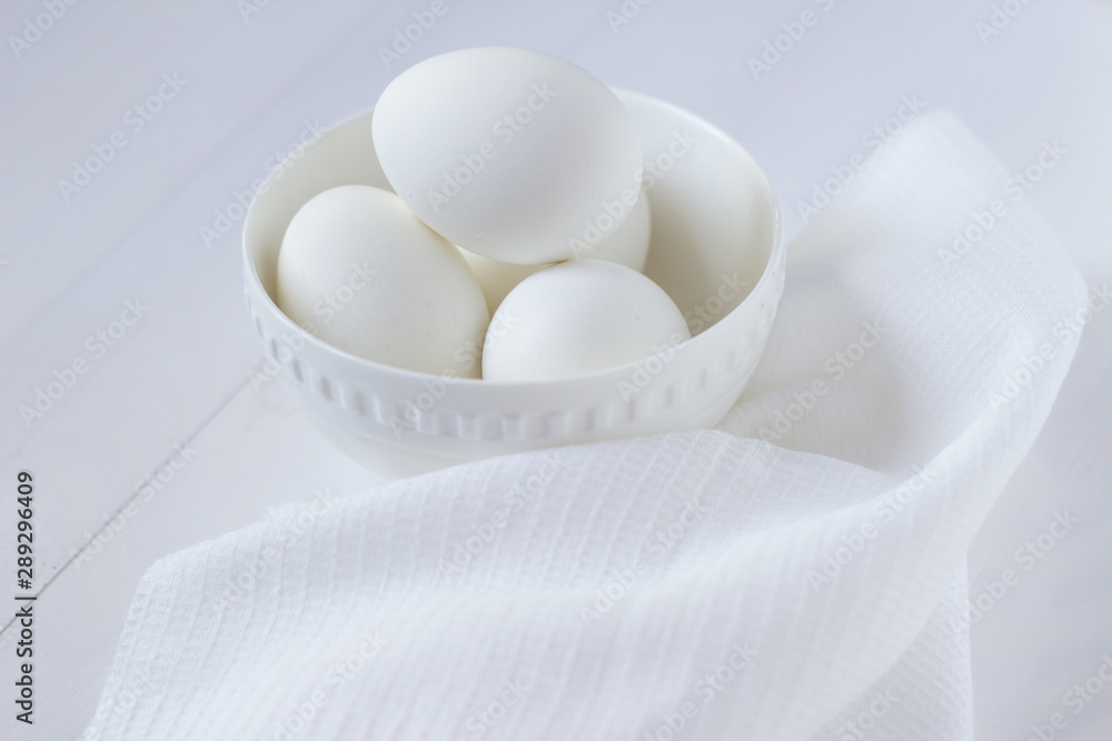 four white eggs lie in a plate on a white wooden table next to a towel. four white eggs lie in a plate on a white wooden table next to a towel.