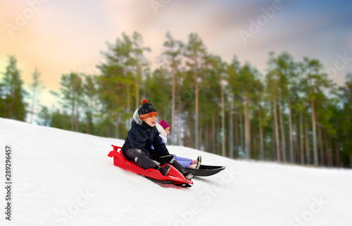 childhood, sledging and season concept - happy little kids sliding on sleds down snow hill over winter forest background