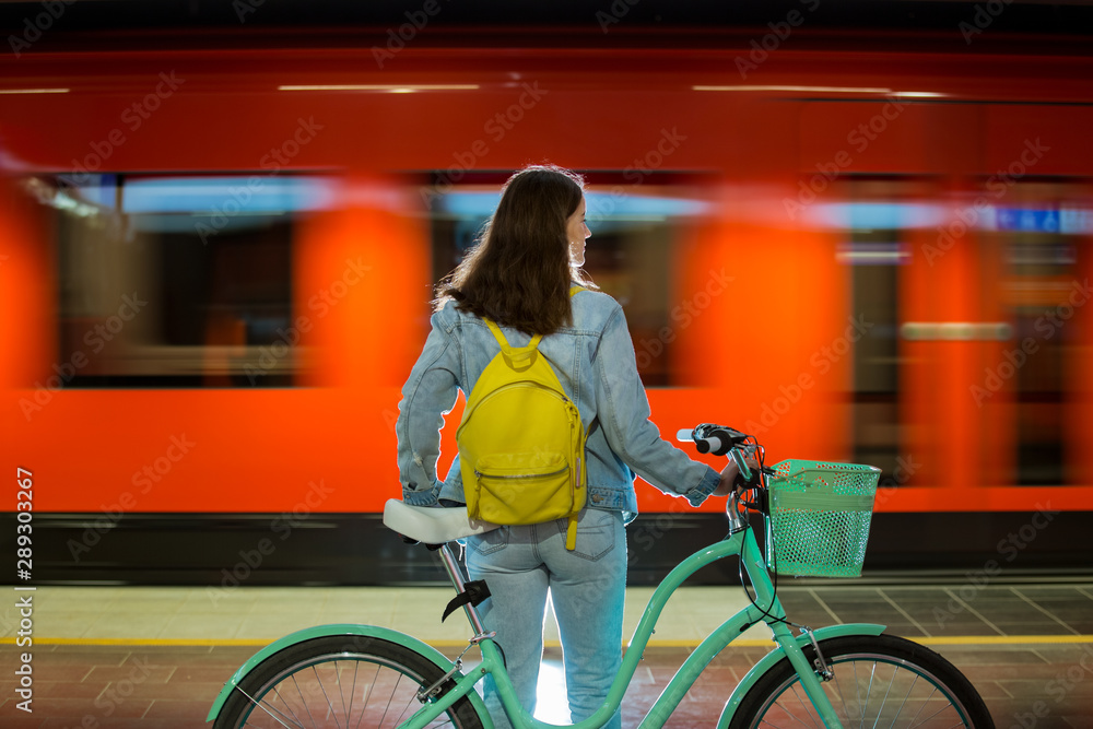 Teenager girl in jeans with yellow backpack and bike standing on metro  station, waiting for train, smiling, laughing. Orange train passing by  behind the girl. Futuristic subway station. Finland, Espoo Stock Photo