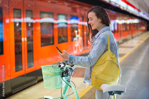 Teenager girl with backpack and bike standing on metro station holding smart phone in hand, scrolling and texting, smiling and laughing. Futuristic bright subway station. Finland, Espoo