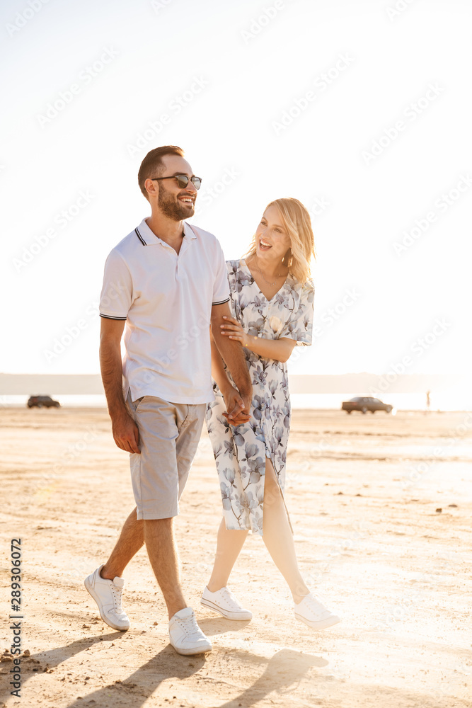 Photo of cheerful beautiful couple laughing and holding hands together while walking on sunny beach Photo of cheerful beautiful couple laughing and holding hands together while walking on sunny beach