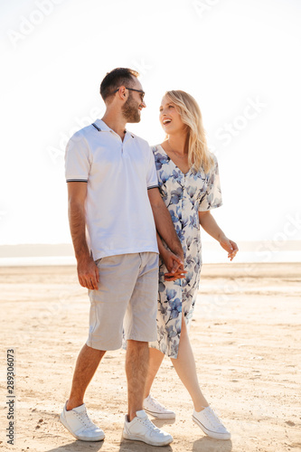 Photo of funny nice couple laughing and holding hands together while walking on sunny beach