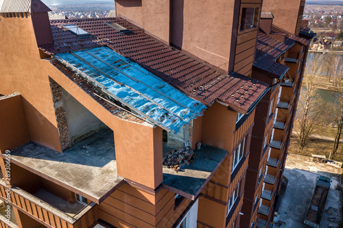 Aerial top view of unfinished tall apartment building, annex room under construction, high chimneys and plastic windows on tiled roof.