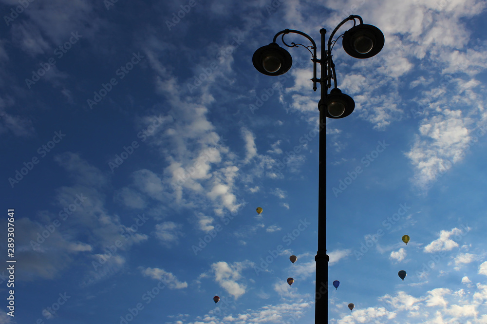 Street lamp pole with blue sky background and hot air balloons seen flying in the distance.