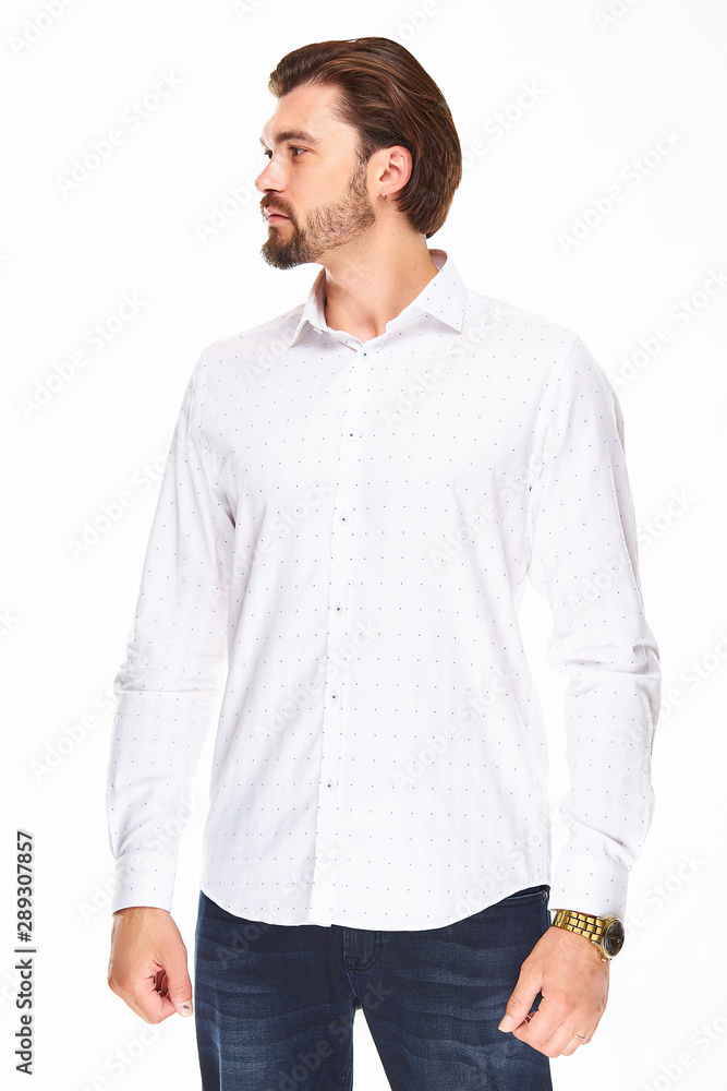 a man with a beard and dark hair stands on a white background in a white shirt in a dot and dark jeans, he has a gold watch on his arm and looks to the left. a man with a beard and dark hair stands on a white background in a white shirt in a dot and dark jeans, he has a gold watch on his arm and looks to the left.