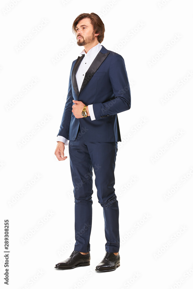 man with beard and dark hair stands on a white background in dark blue tuxedo, white shirt and black leather shoes, he has a gold watch on his arm and he looks away. man with beard and dark hair stands on a white background in dark blue tuxedo, white shirt and black leather shoes, he has a gold watch on his arm and he looks away.