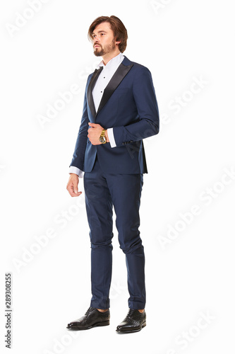 man with beard and dark hair stands on a white background in dark blue tuxedo, white shirt and black leather shoes, he has a gold watch on his arm and he looks away.