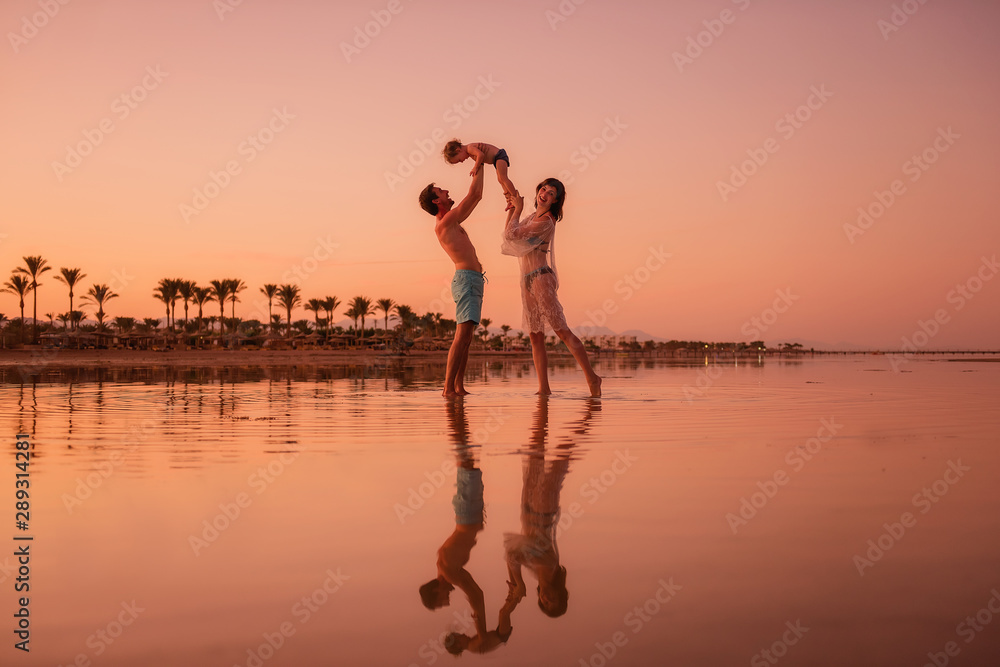 Fototapeta premium A young family travels the world. Parents throw in the arms of a child at sunset. Reflection in water.