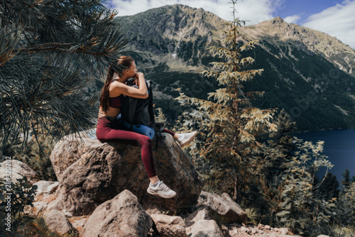 Young attractive girl in red sportswear sitting on the big stone, leaned on her backpack, tired while climbing a mountain, beautiful mountain lake on the background, active lifestyle concept