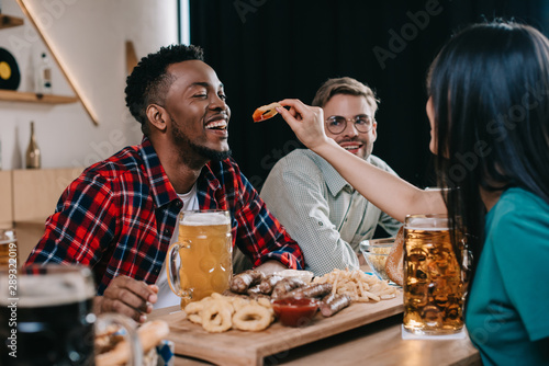 back view of young woman feeding african american man with fried onion ring while celebrating octoberfest in pub