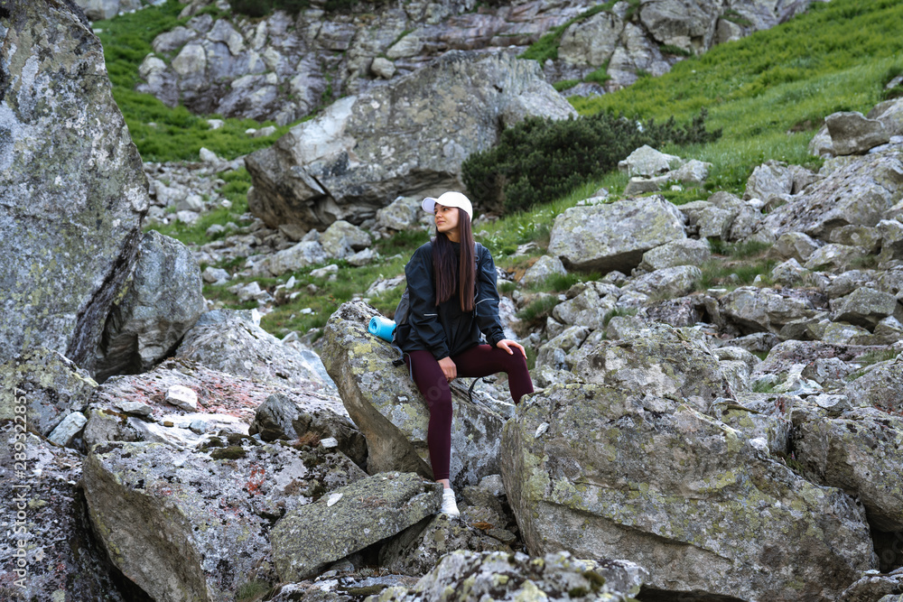 Young woman traveler in white hat and black jacket sitting on big rock among the mountains, active lifestyle concept