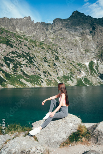Young beautiful sporty girl in grey pants sitting on the big rock and posing on the background of mountain lake, active lifestyle concept