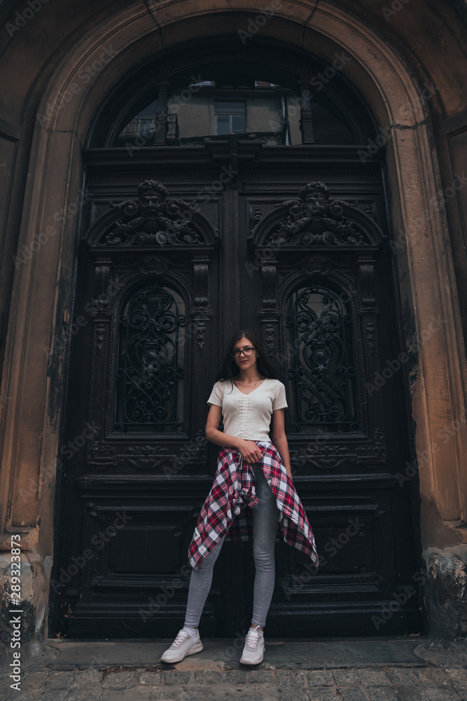 Full length portrait of beautiful young brunette girl in casual clothes and glasses that standing in front of the big old antique black door