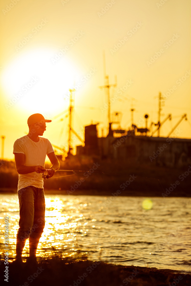 Fototapeta premium Young man fishing at sea from the shore at sunset. soft light. fisherman uses spinning