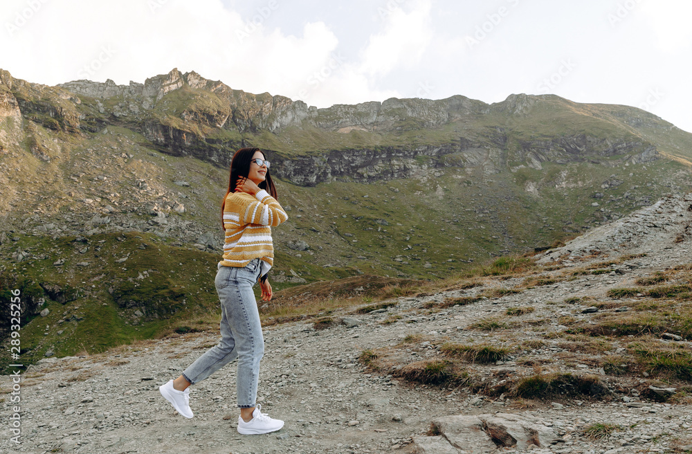 Naklejka premium stylish traveler girl in hat with backpack jumping in mountains. happy hipster woman having fun on top of mountain. space for text. wanderlust and travel concept. atmospheric moment