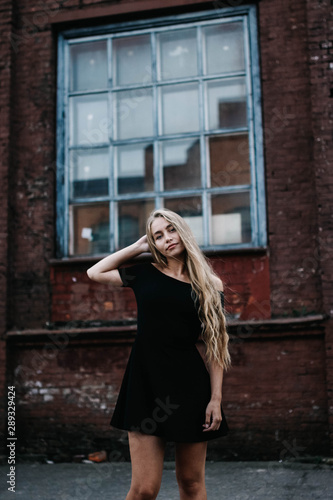 Young blond woman with long hair in a black dress near a brick red wall. Stylish girl in the city.