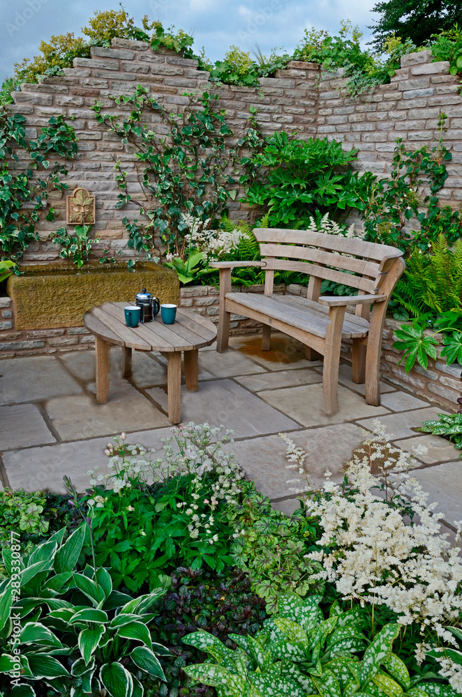 Close up of a wooden garden seat and table in a terrace in a walled garden with seletion of plants
