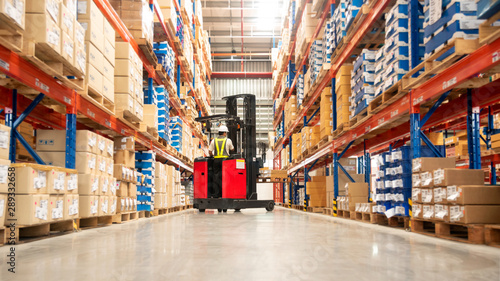 Worker in forklift-truck loading packed goods in huge distribution warehouse with high shelves.