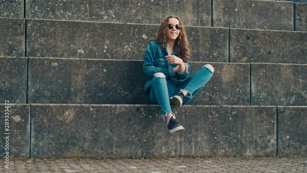 Portrait shot of the young Caucasian stylish hipster girl in jeans clothes and dark sunglasses sitting on the big stoned steps and crossing her legs. Portrait shot of the young Caucasian stylish hipster girl in jeans clothes and dark sunglasses sitting on the big stoned steps and crossing her legs.