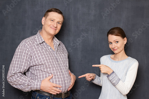 Cheerful woman is pointing at big stomach of satisfied man