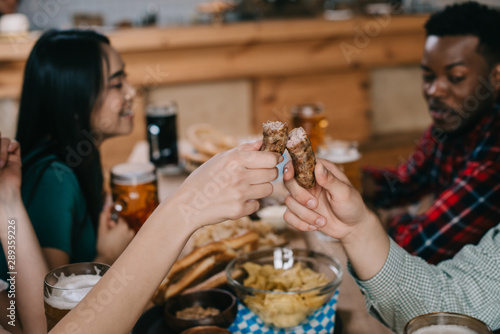 cropped view of young woman and woman holding fried sausage while celebrating octoberfest with multicultural friends