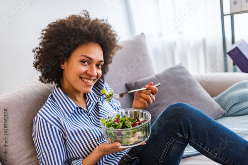 Beautiful young woman having a fresh healthy meal in the living room. Smiling young woman eating salad in the living room at home, healthy food and nutrition concept