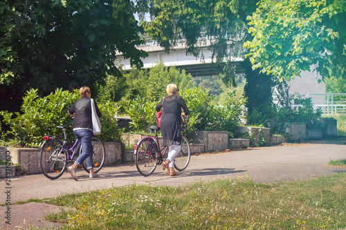 Ladies with bikes in a city park, two middle aged women carry bicycles along the road (unrecognizable people)
