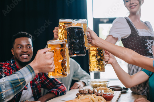 cheerful multicultural friends clinking mugs of beer while celebrating octoberfest in pub