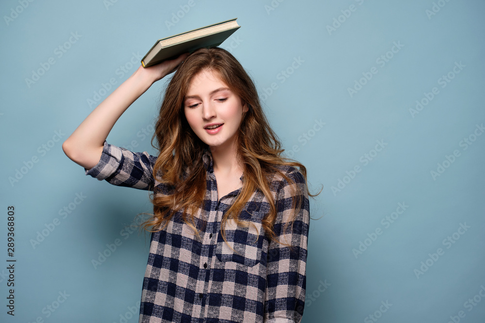 A young girl in a plaid shirt stands on a blue background and looks down with a book on her head. A young girl in a plaid shirt stands on a blue background and looks down with a book on her head.