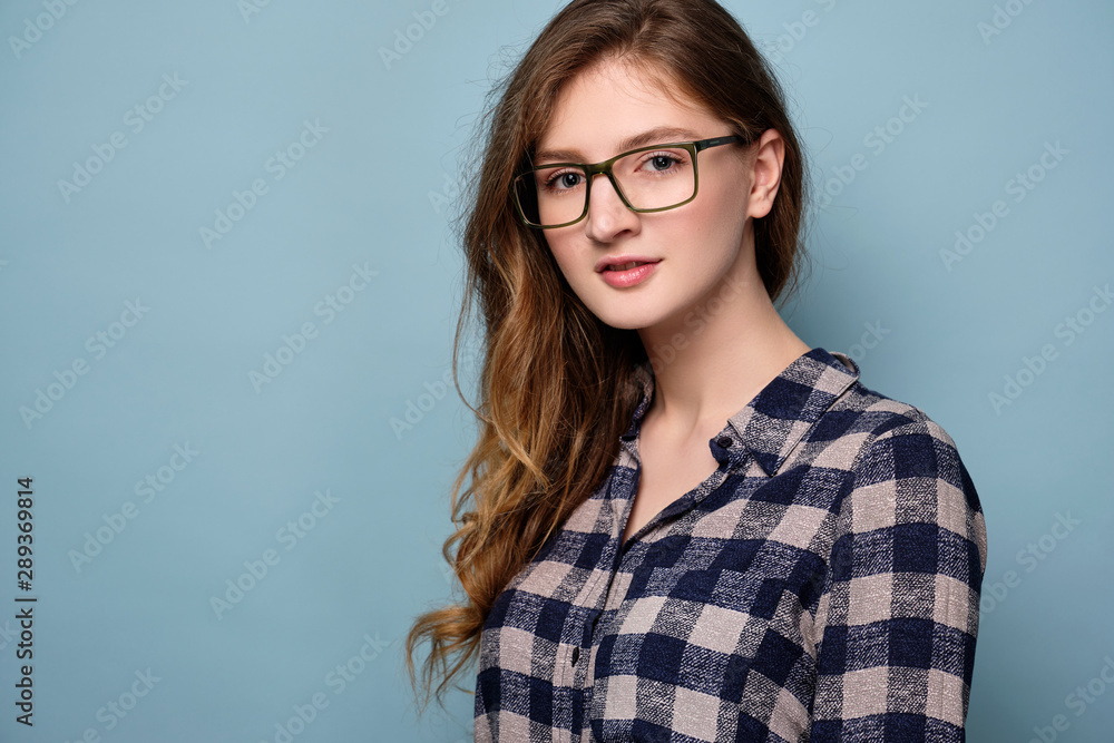 A young girl in a plaid dress and glasses stands on a blue background and looks at the camera. A young girl in a plaid dress and glasses stands on a blue background and looks at the camera.