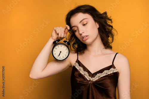 A curly dark-haired girl stands on a yellow background in a brown top and sleepily looks at the alarm clock in her hand.