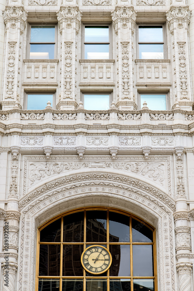 Ornate white facade of an historic building with a clock with Roman numerals and a large window reflecting modern office buildings - old building in downtown Chicago