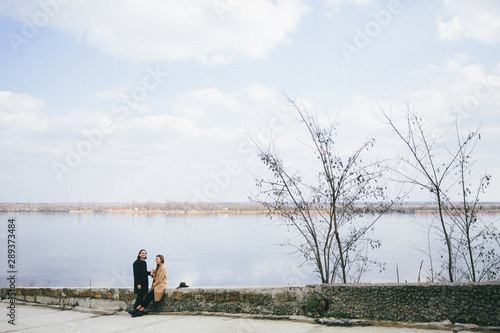 young romantic couple in coats enjoying autumn landscape on the river background