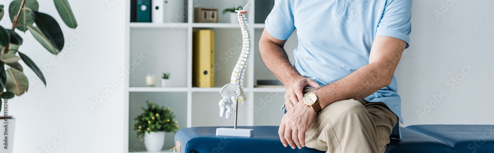 panoramic shot of man sitting on massage table near spine model