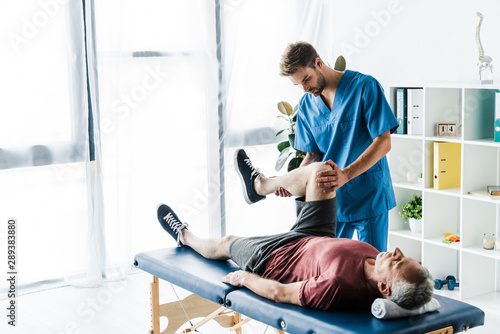 handsome doctor touching leg of mature patient lying on massage table