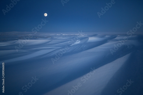 Winter arctic landscape. Evening twilight. Full moon over the snow-covered tundra. A strong wind had formed ridges (sastrugi) on the surface of the snow. Cold polar climate. Chukotka, Siberia, Russia.