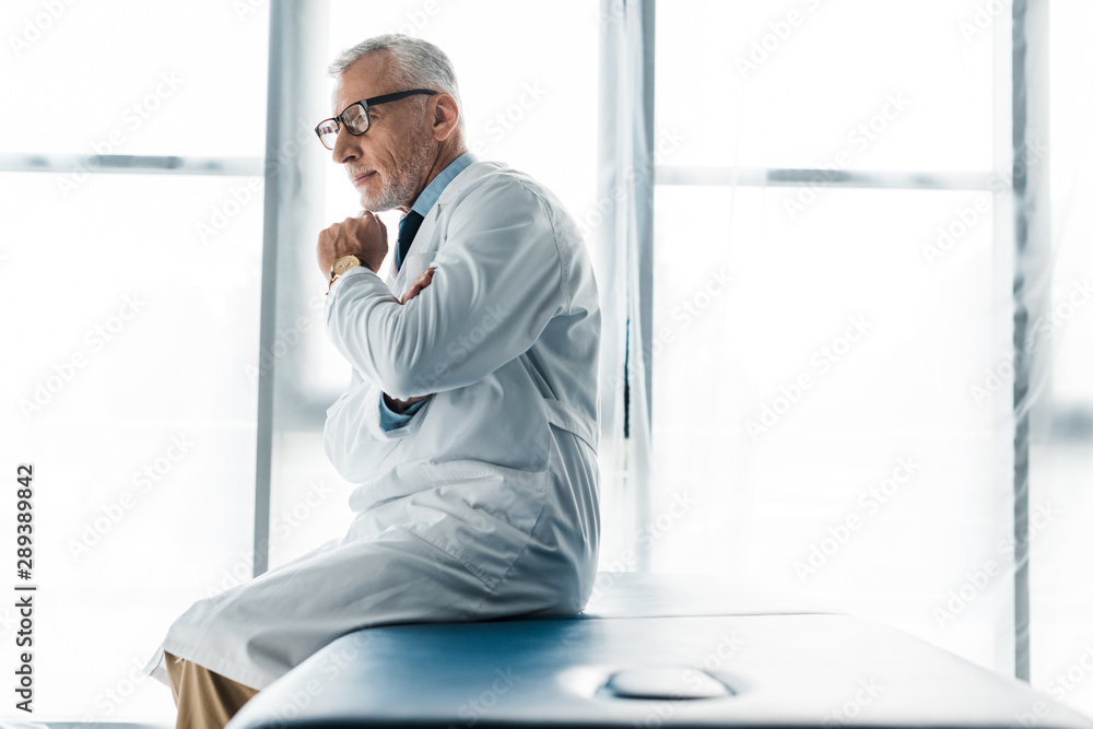 Fototapeta premium thoughtful doctor in glasses and white coat sitting on massage table and touching face