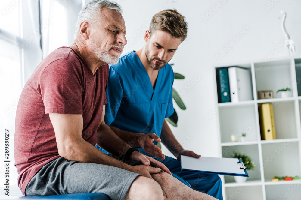 bearded mature patient with closed eyes touching leg near doctor holding clipboard in clinic