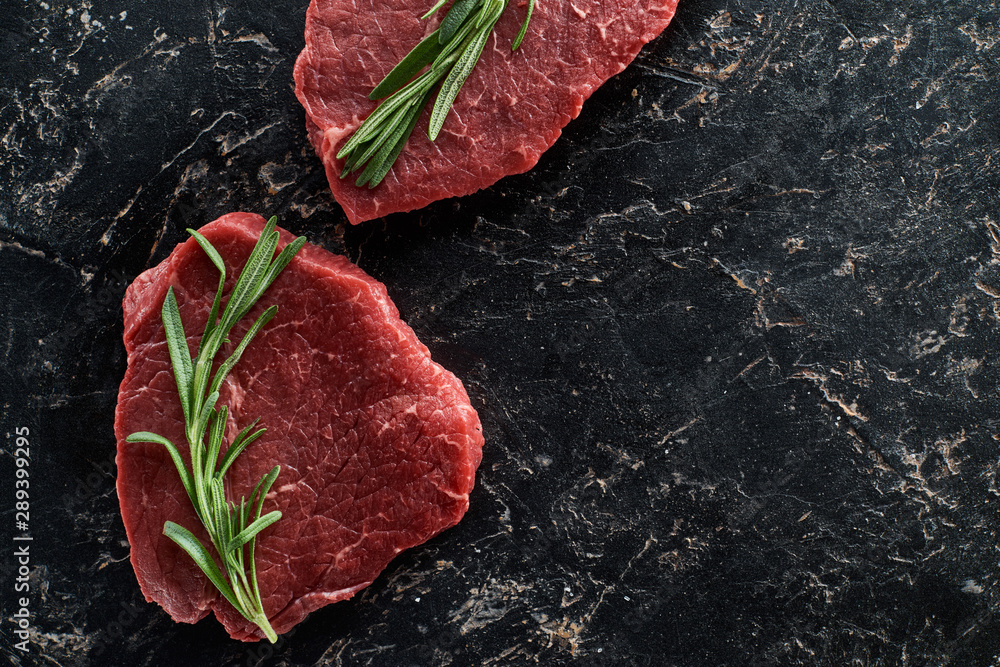top view of uncooked beef sirloins with rosemary on black marble background
