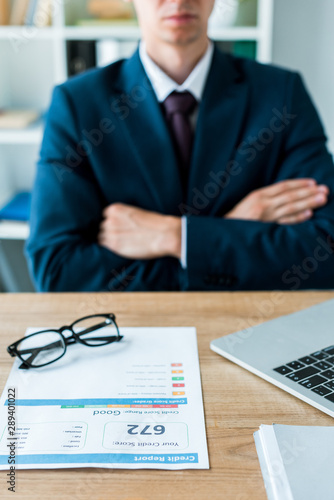 selective focus of document with credit report near laptop and man with crossed arms