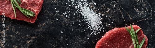 panoramic shot of panoramic shot of raw beef steaks with rosemary swig near scattered salt on black surface