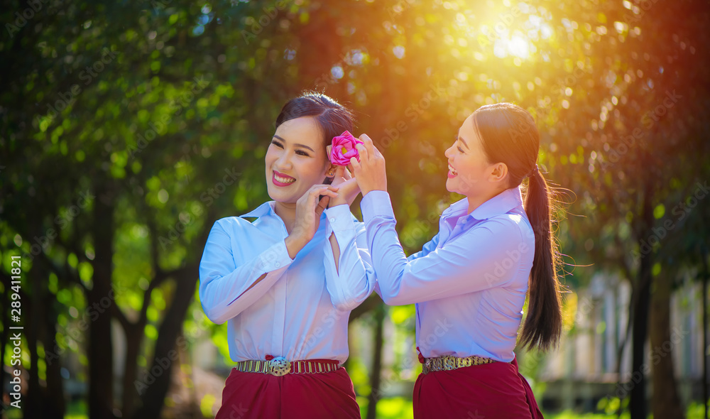 Obraz premium Two beautiful asian women friends in white shirt laughing and holding pink lotus flowers. Two young asian women with sarong cloth decorated with silver belt according to Thai culture, Thailand.